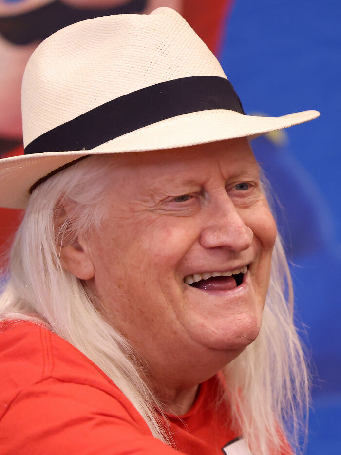 A close-up of Charles Martinet smiling and laughing, wearing a white fedora and a red shirt, showcasing his career highlights.