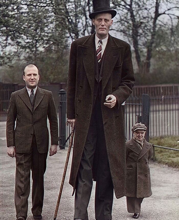 Historical colorized photo featuring a very tall man in a top hat and coat walking with two other men.