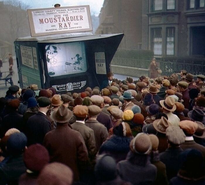 Historical colorized photo of a crowd watching Mickey Mouse on a screen, making the past days come to life.