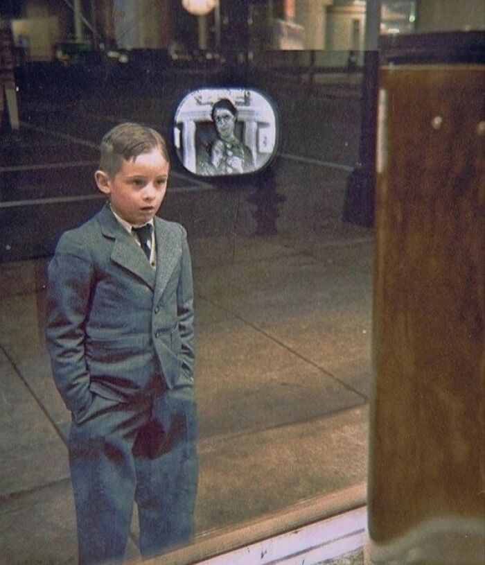 A historical colorized photo depicts a young boy looking into a store window with a reflection, bringing past days to life.