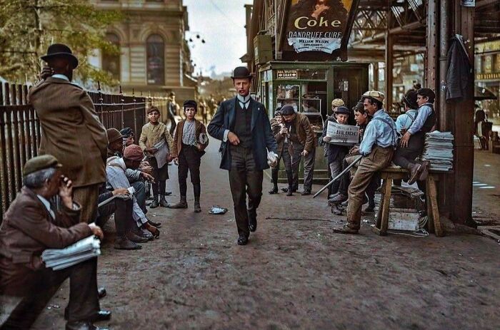 Historical colorized photo of a busy street scene with people and shops.