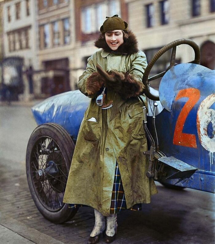 A historical colorized photo of a woman in a long coat standing next to a vintage race car. Stunning historical colorized photos.