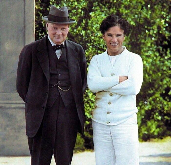 Winston Churchill in a suit and hat stands next to Charlie Chaplin in white attire, captured in a historical colorized photo.