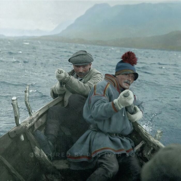 A stunning historical colorized photo of two men rowing a boat in choppy water with mountains in the background.