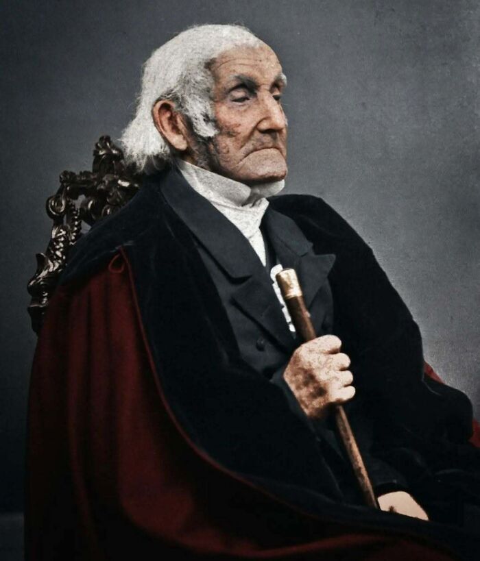 A historical colorized photo of an old man in a suit holding a cane and sitting on a chair. Stunning historical colorized photos.