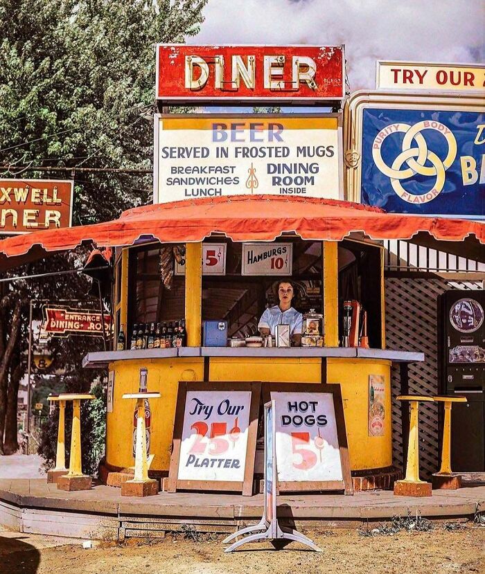Historical colorized photo of a vibrant vintage diner stand with a person inside and colorful signs.