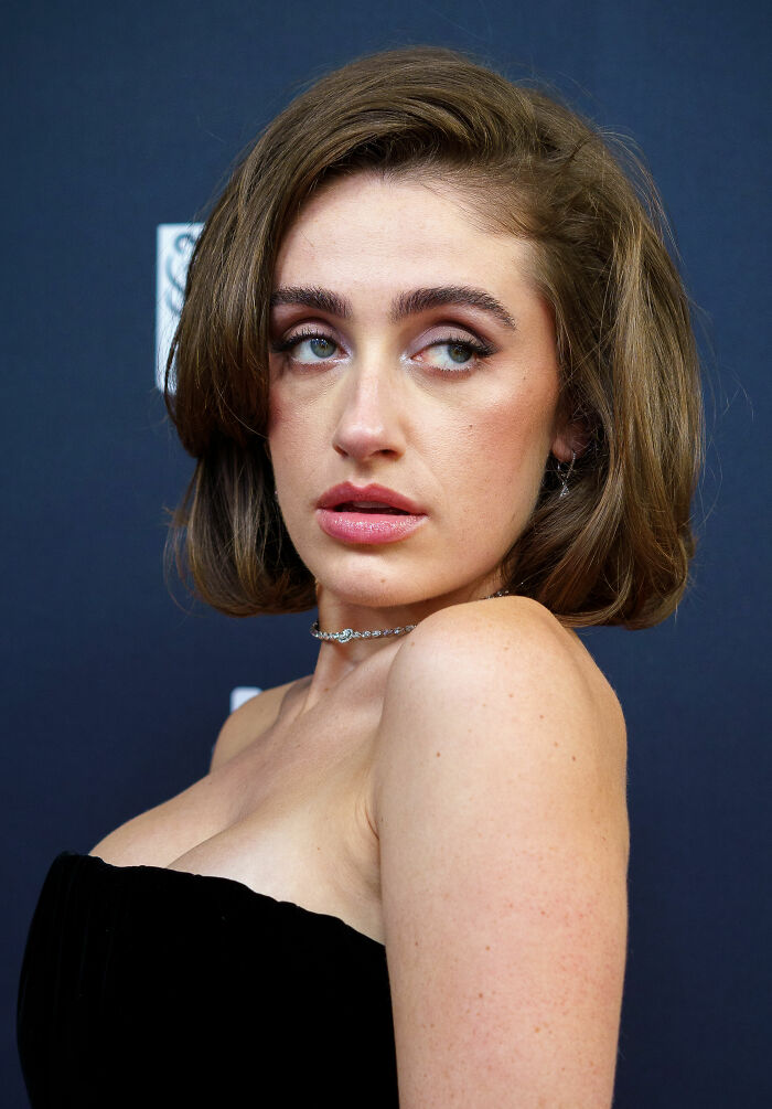 A close-up of Rachel Sennott's face, highlighting her career highlights and bio. Her short brown hair frames her face, with soft makeup emphasizing her eyes and lips. She wears a strapless black top and a delicate choker necklace against a dark background, reflecting her rising career.
