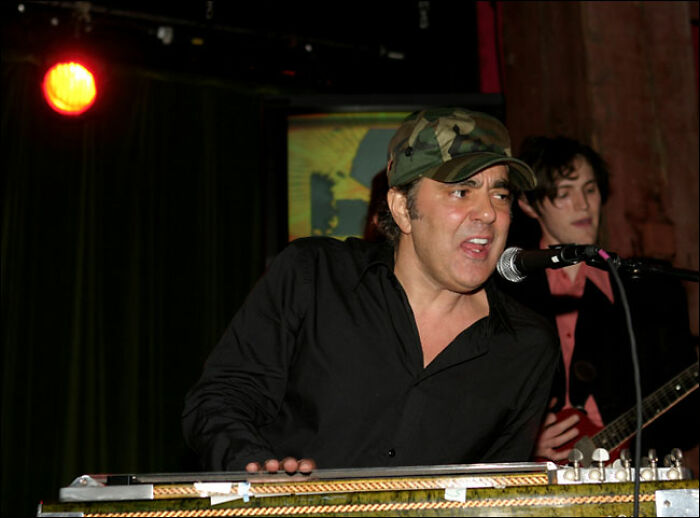 Daniel Lanois performing on stage, wearing a camouflage hat and black shirt, playing an instrument with a microphone in front of him, showcasing his career highlights.