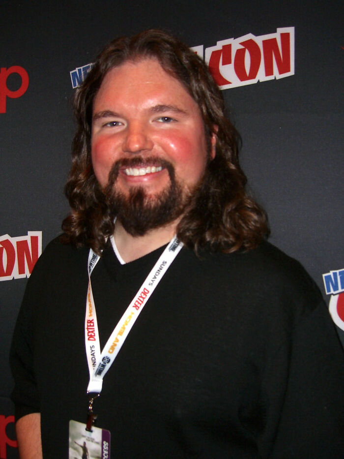 A smiling Marcus Dunstan, with long hair and a beard, wearing a black shirt and a lanyard, poses for a photo.