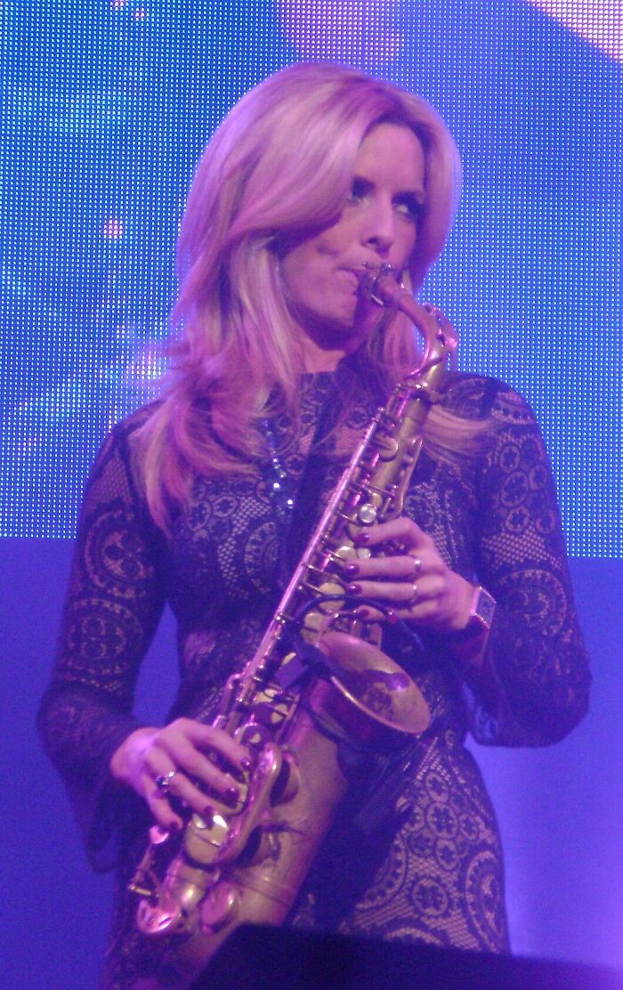 Candy Dulfer performing live, playing a saxophone with focus. She wears a black lace dress, illuminated by blue stage lights.