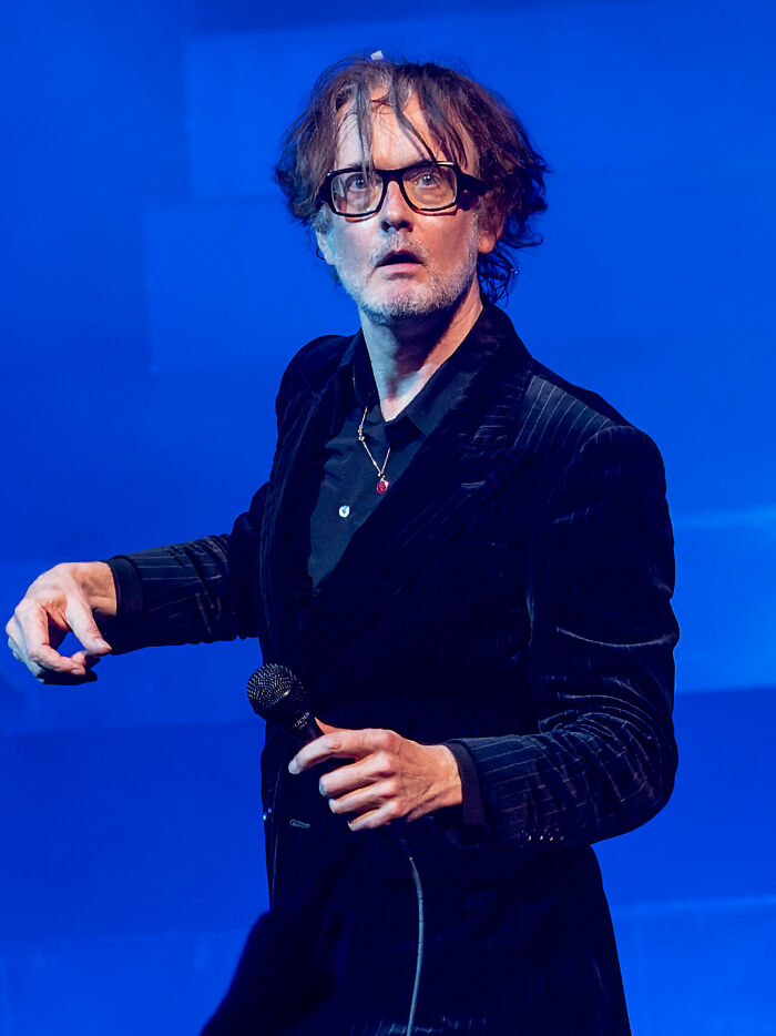 Jarvis Cocker performs live, wearing glasses and holding a microphone, showcasing his iconic career highlights.