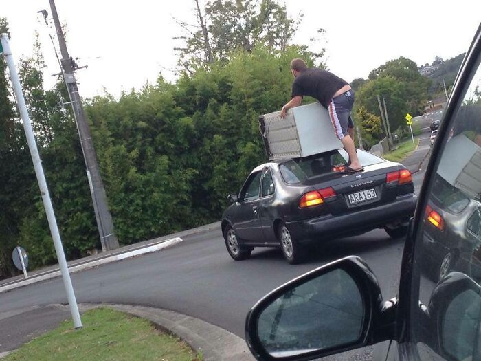 Man standing on car roof holding a large appliance, a risky moving fails activity.