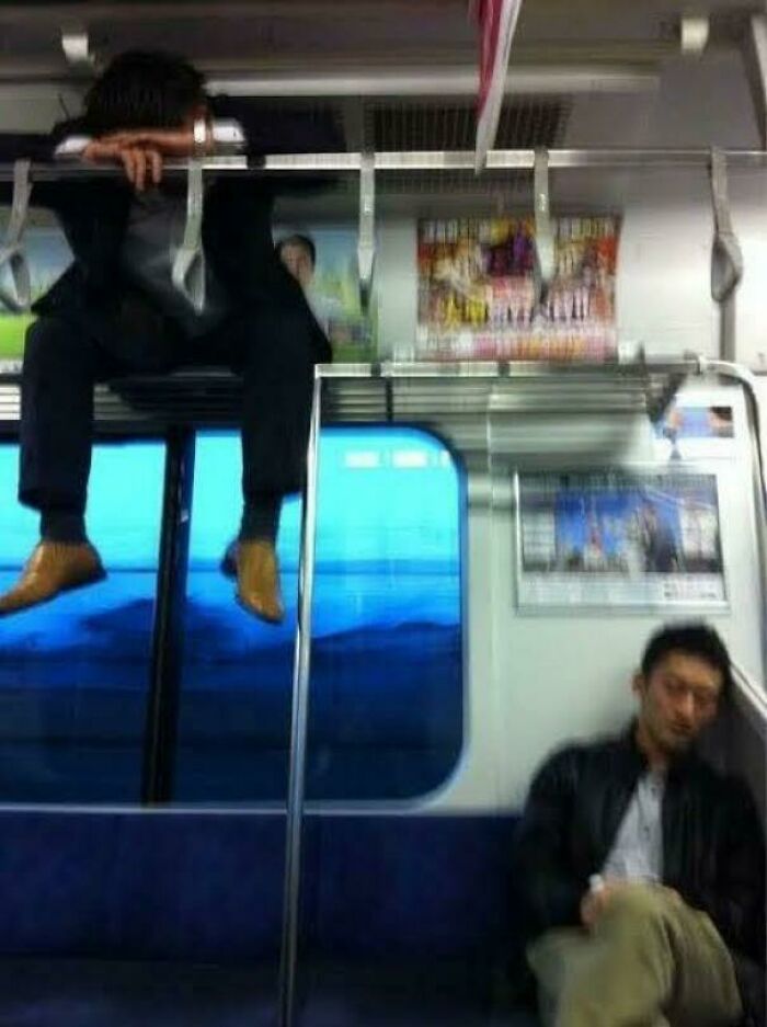 A man sleeping precariously on the overhead luggage rack during public transport travel