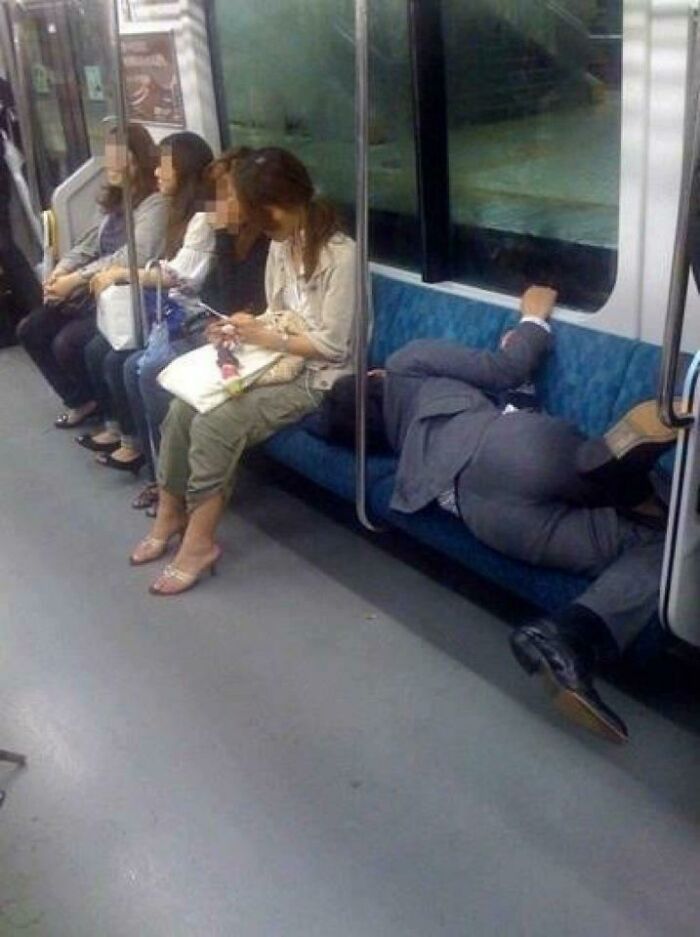 A tired man sleeping horizontally on public transport subway seats while other passengers sit nearby