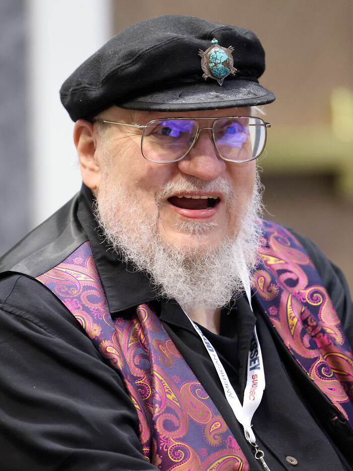 Author George R. R. Martin smiling at an event wearing his signature black cap and glasses.