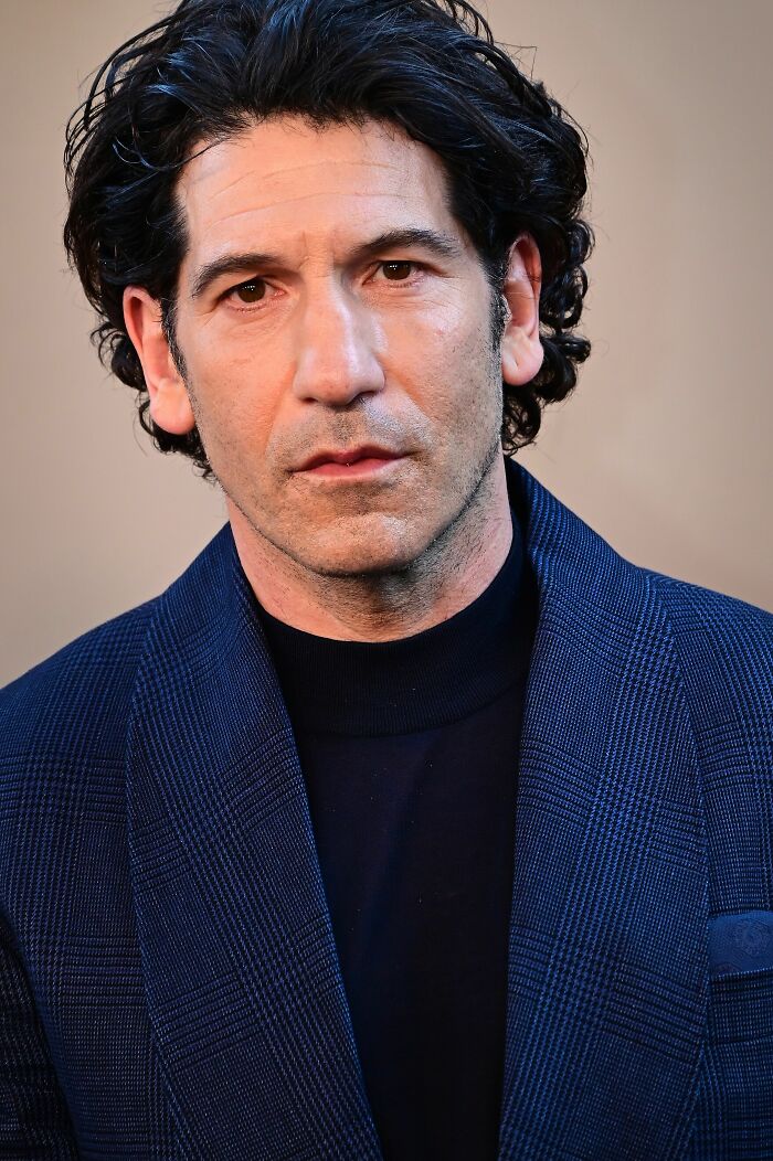 A close-up portrait of actor Jon Bernthal wearing a blue plaid suit jacket over a dark turtleneck.