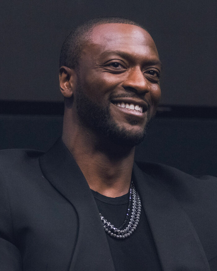 A close-up portrait of actor Aldis Hodge smiling in a black suit jacket and necklace