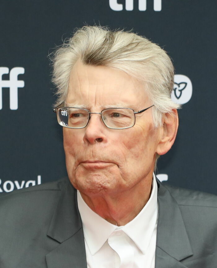 A close-up portrait of author Stephen King wearing glasses and a grey suit jacket.