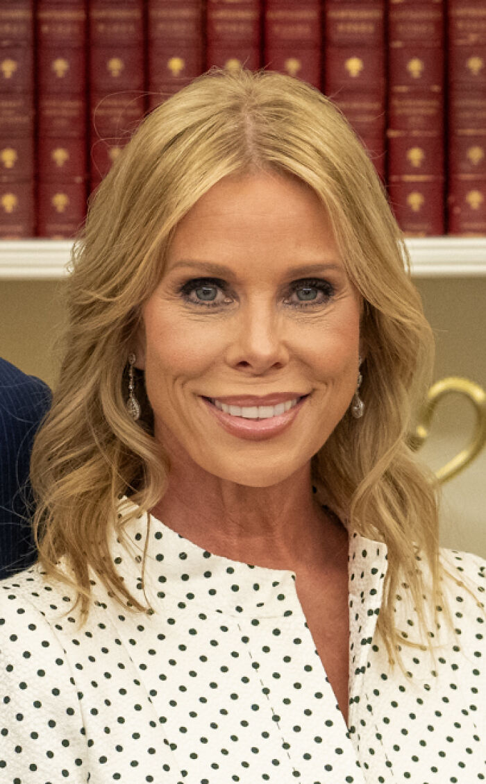 A close-up portrait of Cheryl Hines smiling in a white polka-dot dress with a bookshelf in the background.