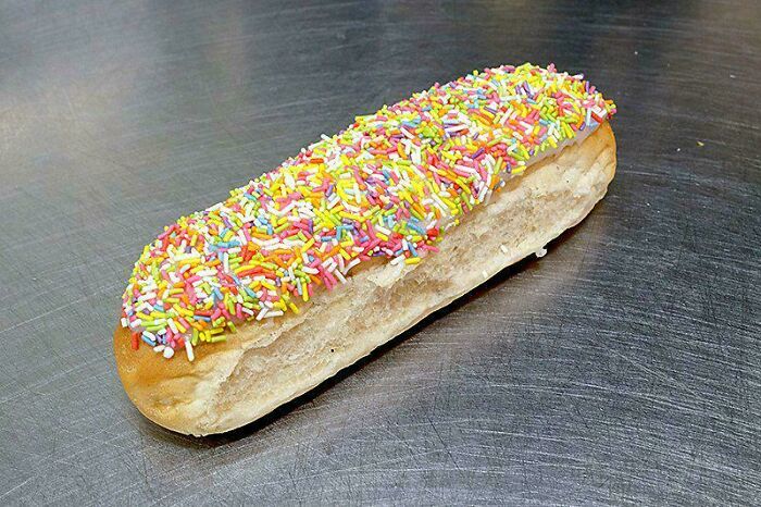 A long bun covered in colorful sprinkles on a metal table, illustrating relatable British memes about bakery treats.