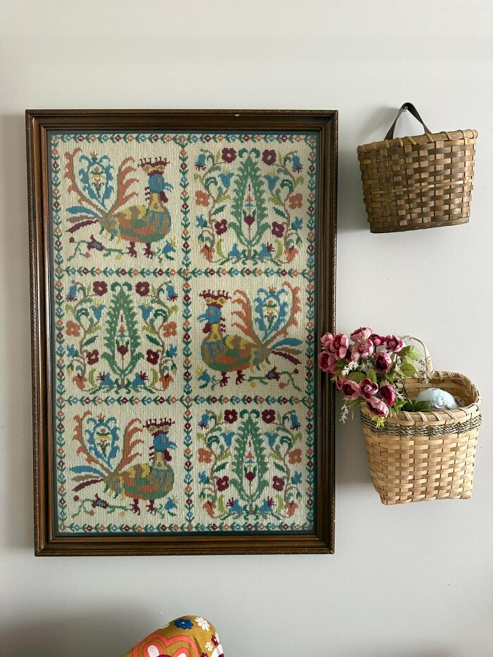 A framed tapestry with rooster designs hung next to small baskets, representing unique thrift store finds