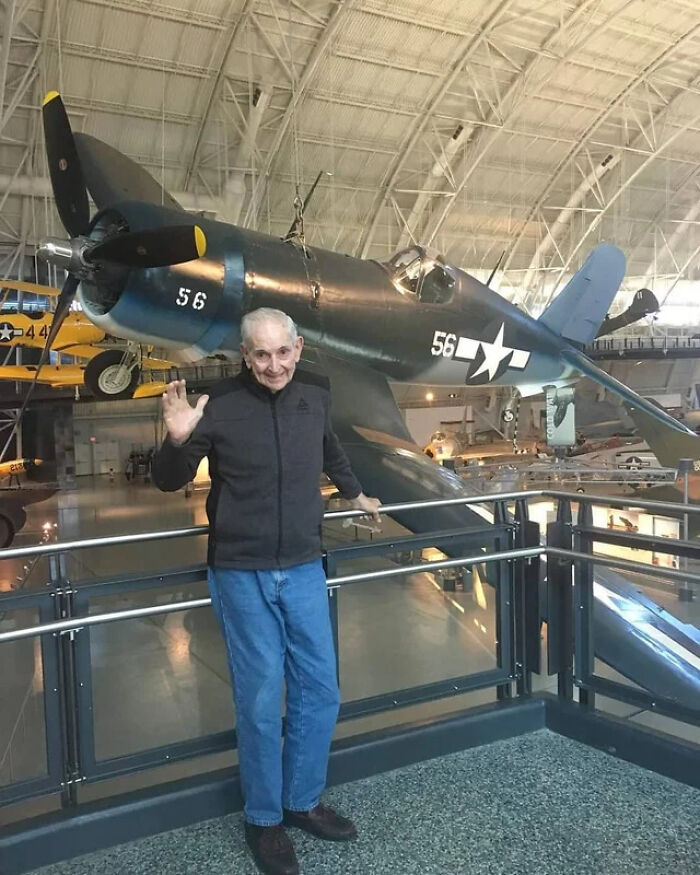 An interesting photo of an elderly man waving in front of a vintage military airplane.
