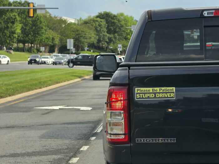 A black pickup truck featuring wild bumper stickers that read Please Be Patient STUPID DRIVER on its tailgate.
