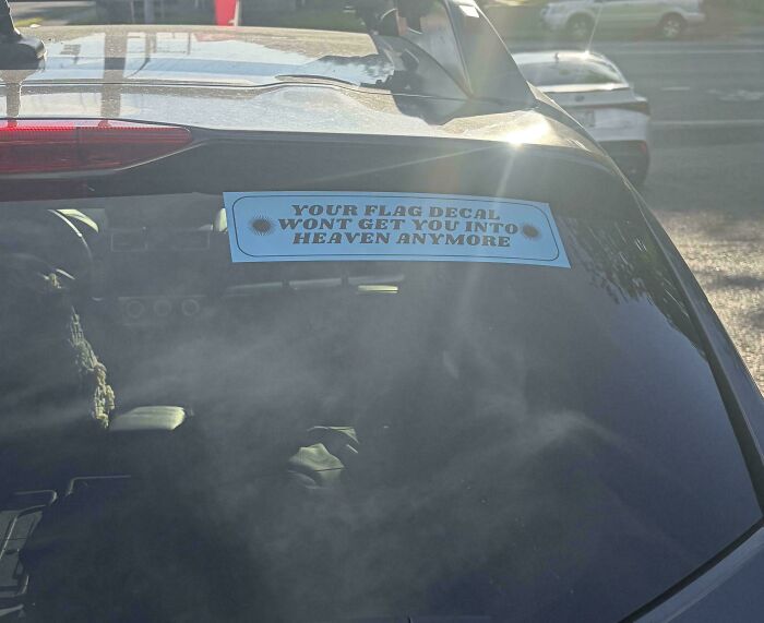 A car rear window displaying one of many wild bumper stickers that reads Your Flag Decal Won't Get You Into Heaven.