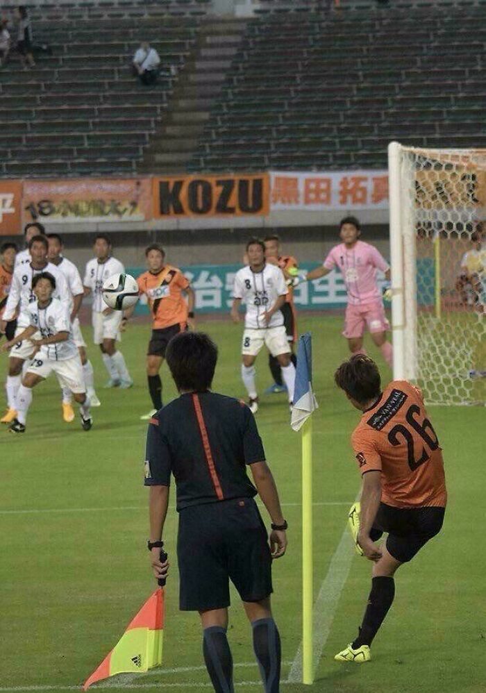 Soccer player kicks ball near corner flag while referee and players watch, creating an optical illusion