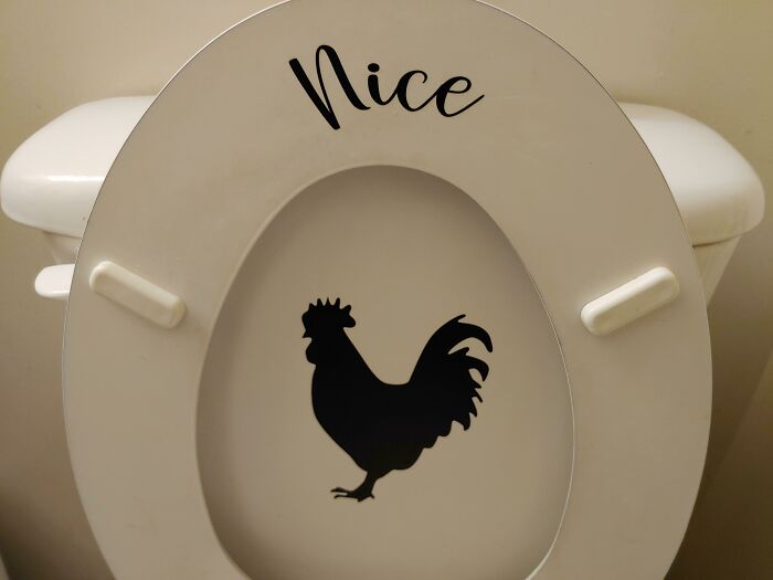 A white toilet seat with a black rooster decal underneath a sticker that reads Nice