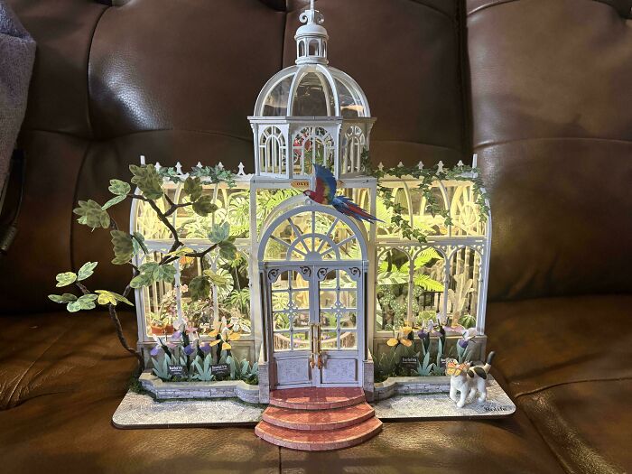 A glass greenhouse book nook miniature featuring a parrot and green plants inside