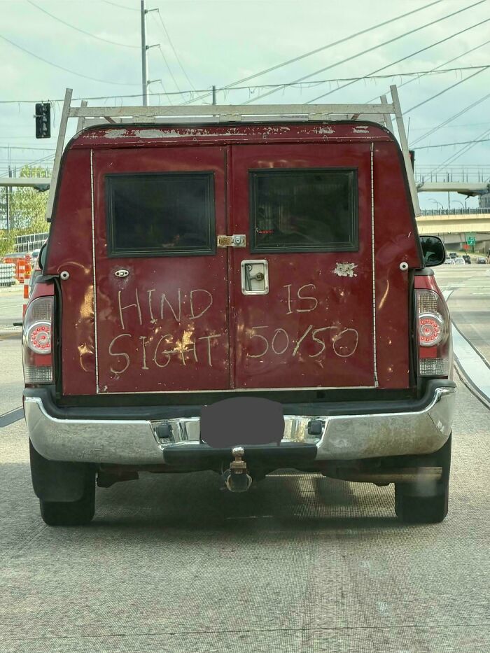 Hand-scratched text on a red truck door serving as one of many badly designed signs reading HINDSIGHT IS 50/50.