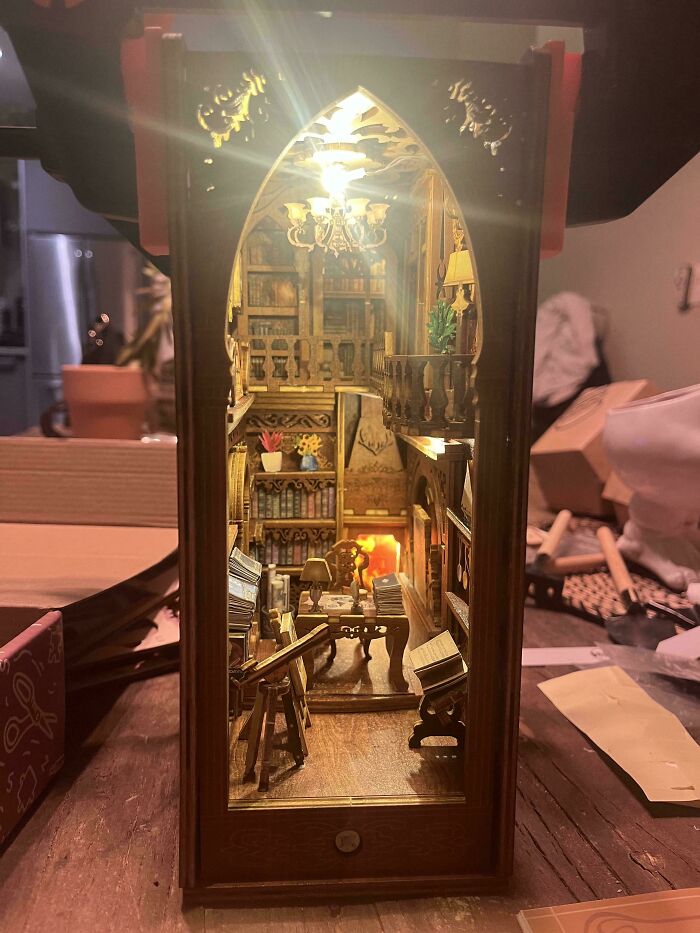 A glowing, detailed library book nook with miniature bookshelves, an armchair, and a tiny chandelier.