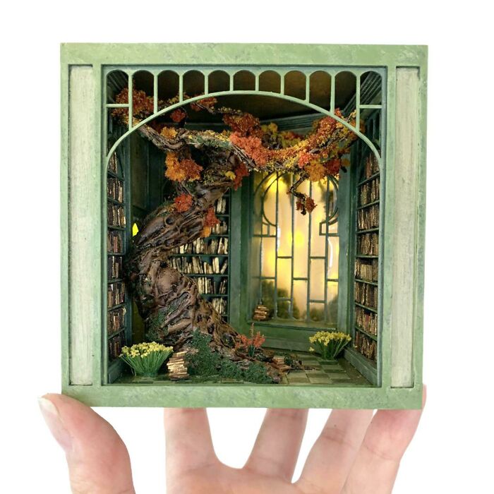 Hand holding a mini green book nook with a tiny tree, bookshelves, and glowing window.