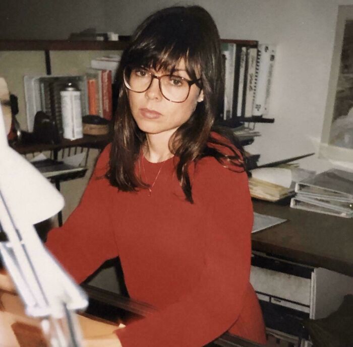 A young woman wearing 90s style large round glasses and a red sweater sitting at a desk