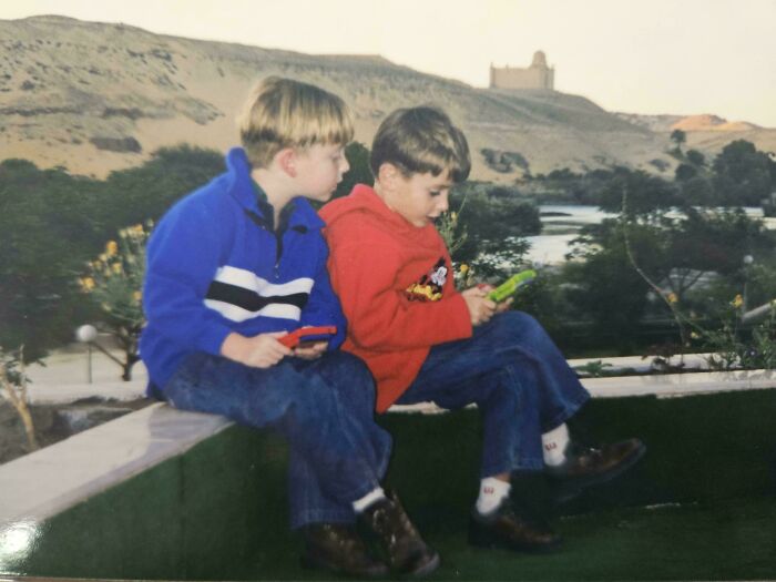 Two boys sitting outdoors playing on their Game Boy handheld consoles, reliving the '90s era.