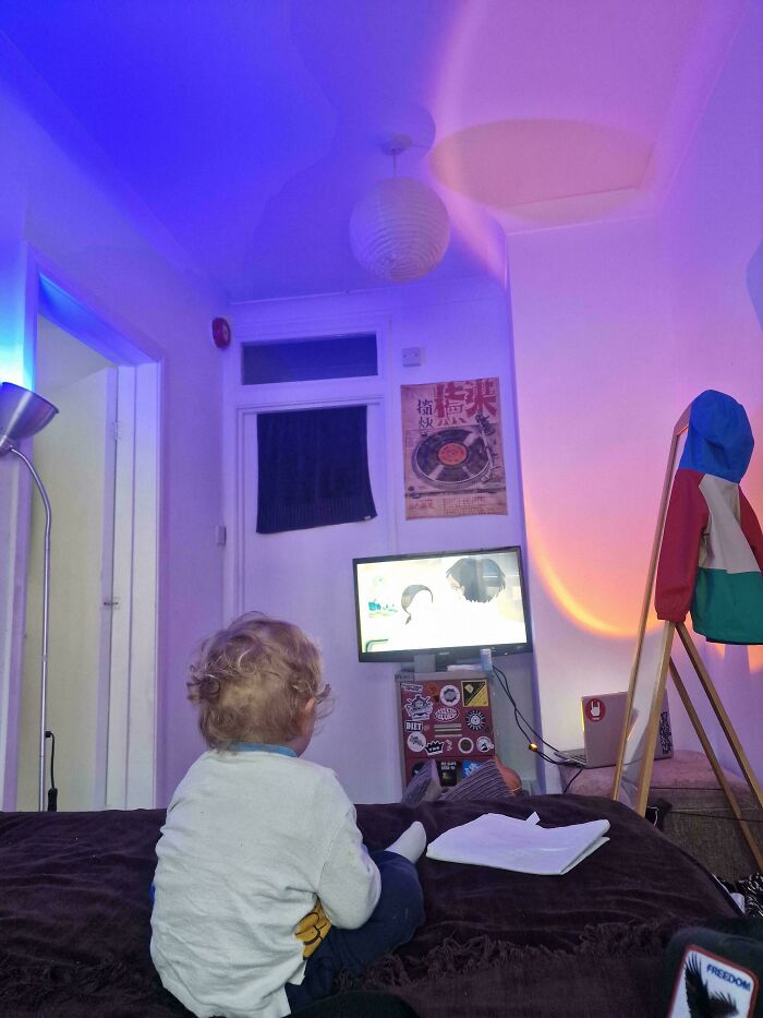 A toddler watching television in a bedroom illuminated by colorful mood lighting, from wholesome pics of dads.