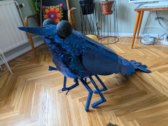 A blue sequined bird sculpture standing indoors, an awful creation far too well-made to ignore.