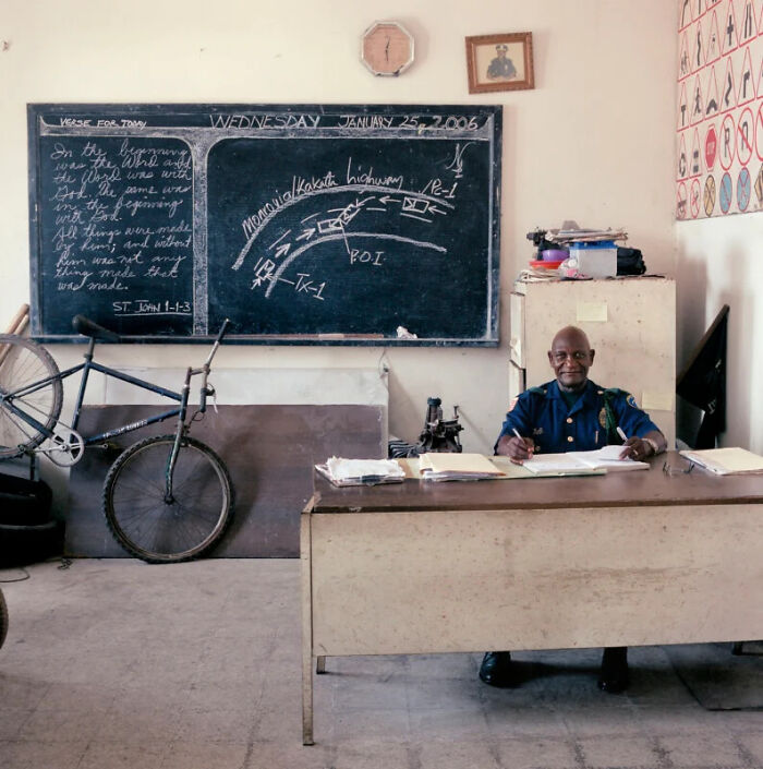 A man in uniform sits at a desk with a chalkboard behind him, documenting the people and offices behind bureaucracy.