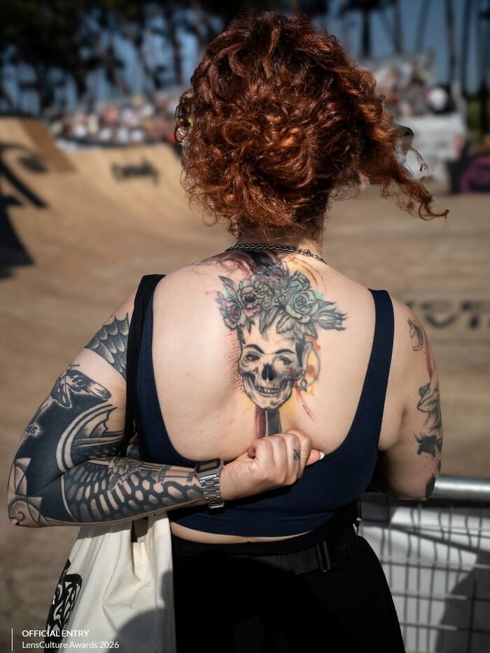 A street photographer captures an odd scene: a woman with numerous tattoos on her back and arms at a skate park.