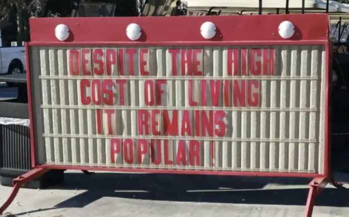 A chaotic sign in the wild that says Despite The High Cost Of Living, It Remains Popular! on a red display.