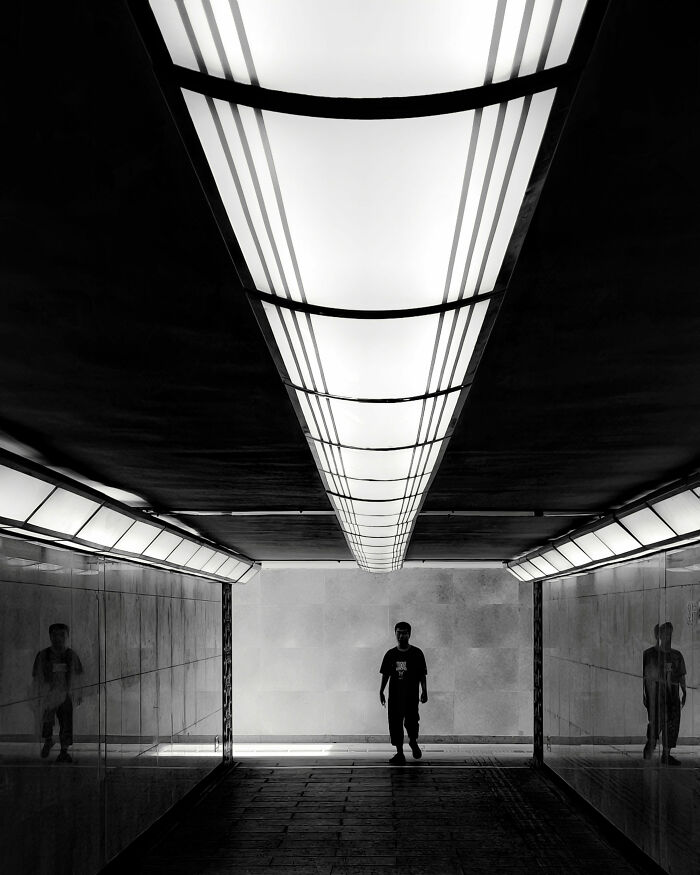 A bold minimalist scene in black and white, featuring a person walking under a long, illuminated architectural structure.