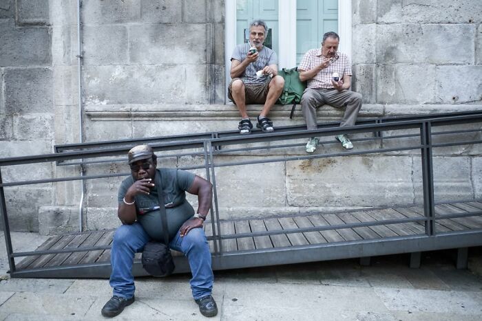 A street photographer captures three men relaxing on a street, showcasing a scene people walk past.