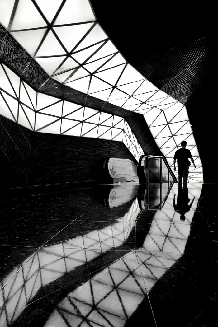 Monochromatic architecture photography showing a person near an escalator with geometric ceiling lights and reflections, a minimalist scene.
