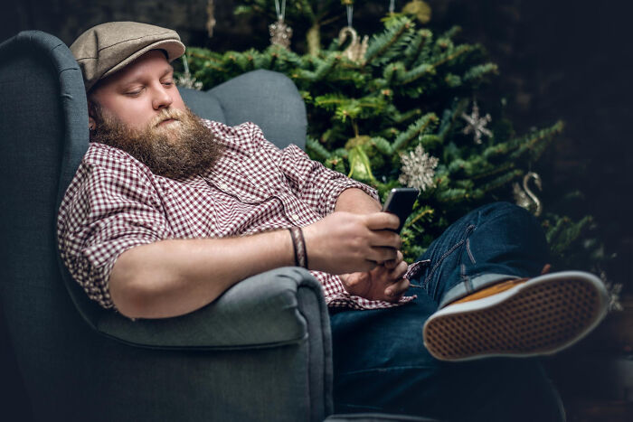 Bearded man in a hat relaxing in a chair looking at his phone. Represents waiting or dealing with a pure nightmare seatmate.