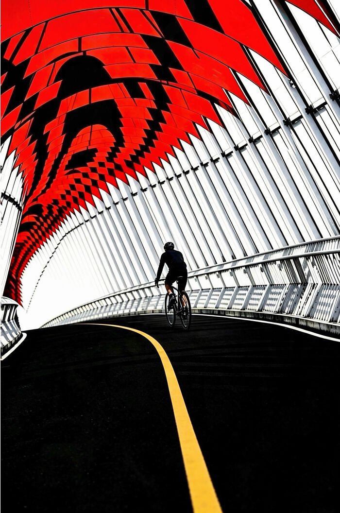 Bold minimalist scene of a cyclist on a curving path inside a striking architectural structure with red patterns.