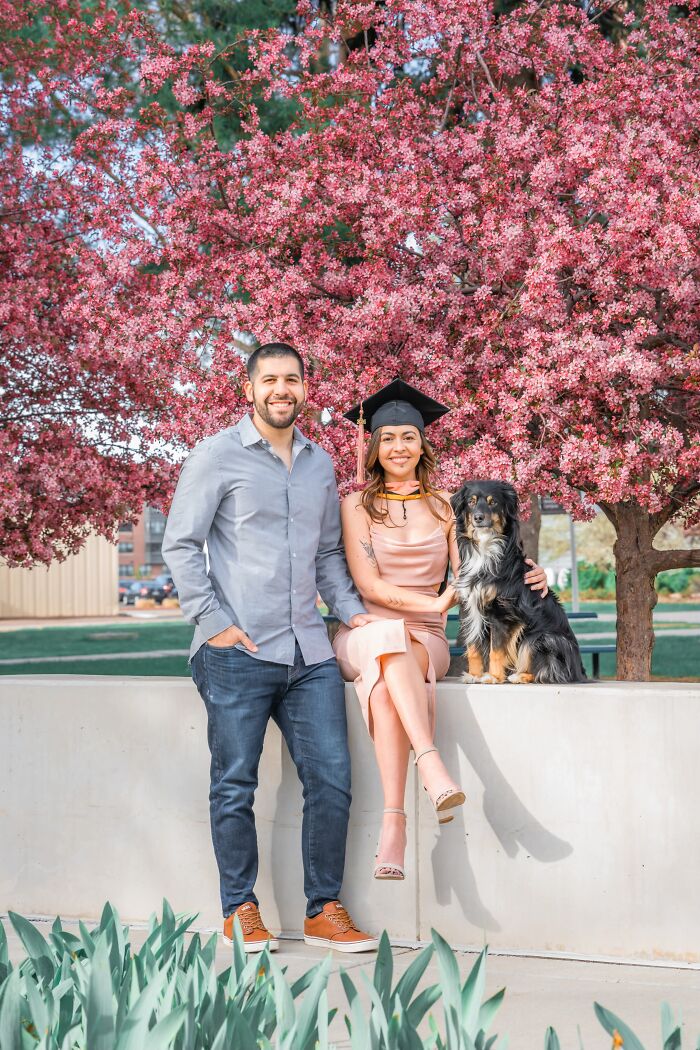 A couple and their dog sitting under a blossoming tree, a woman in a graduation cap, showing sweet proof of finding the right man.