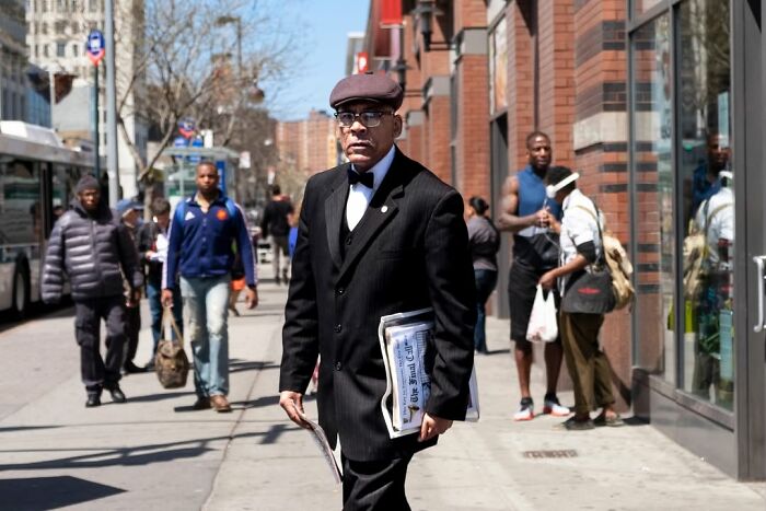 A street photographer captures an odd coincidence: a well-dressed man walks on a sunny street, carrying newspapers.