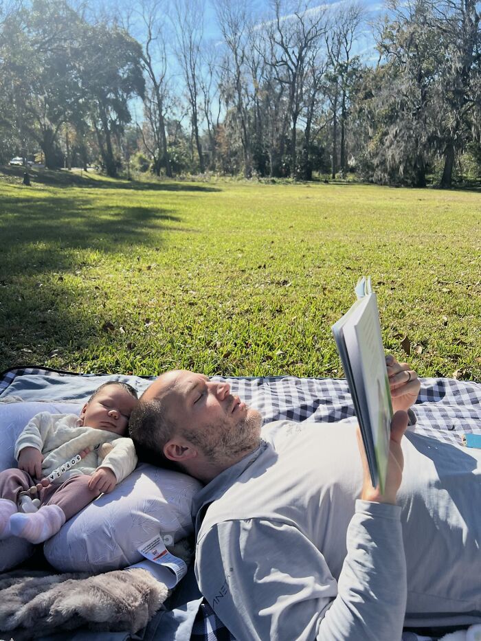 A father relaxing in a park, reading a book while his baby naps beside him, a sweet proof of finding the right man for a family.