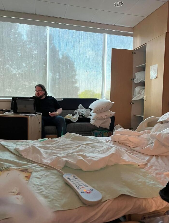 A man sitting on a sofa in a hospital room, working on a laptop, proof of the right man.